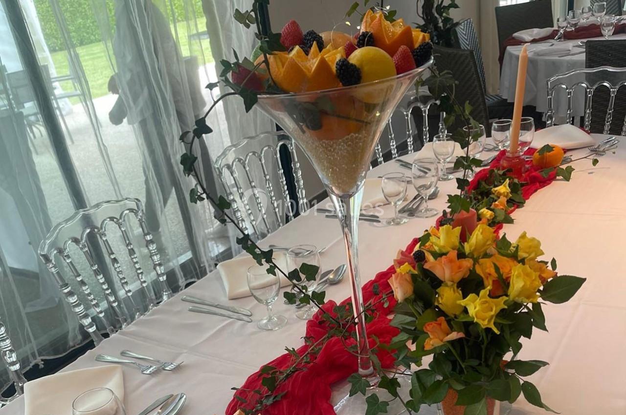 Table décorée avec une grande coupe de fruits, des fleurs colorées et une nappe blanche ornée d'un chemin de table rouge.