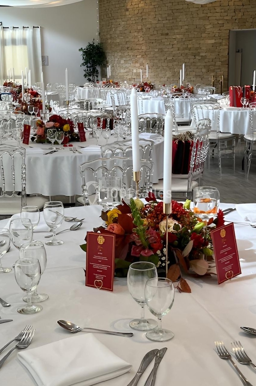 Salle de réception décorée avec des tables rondes, nappes blanches, chaises transparentes et centres de table fleuris.