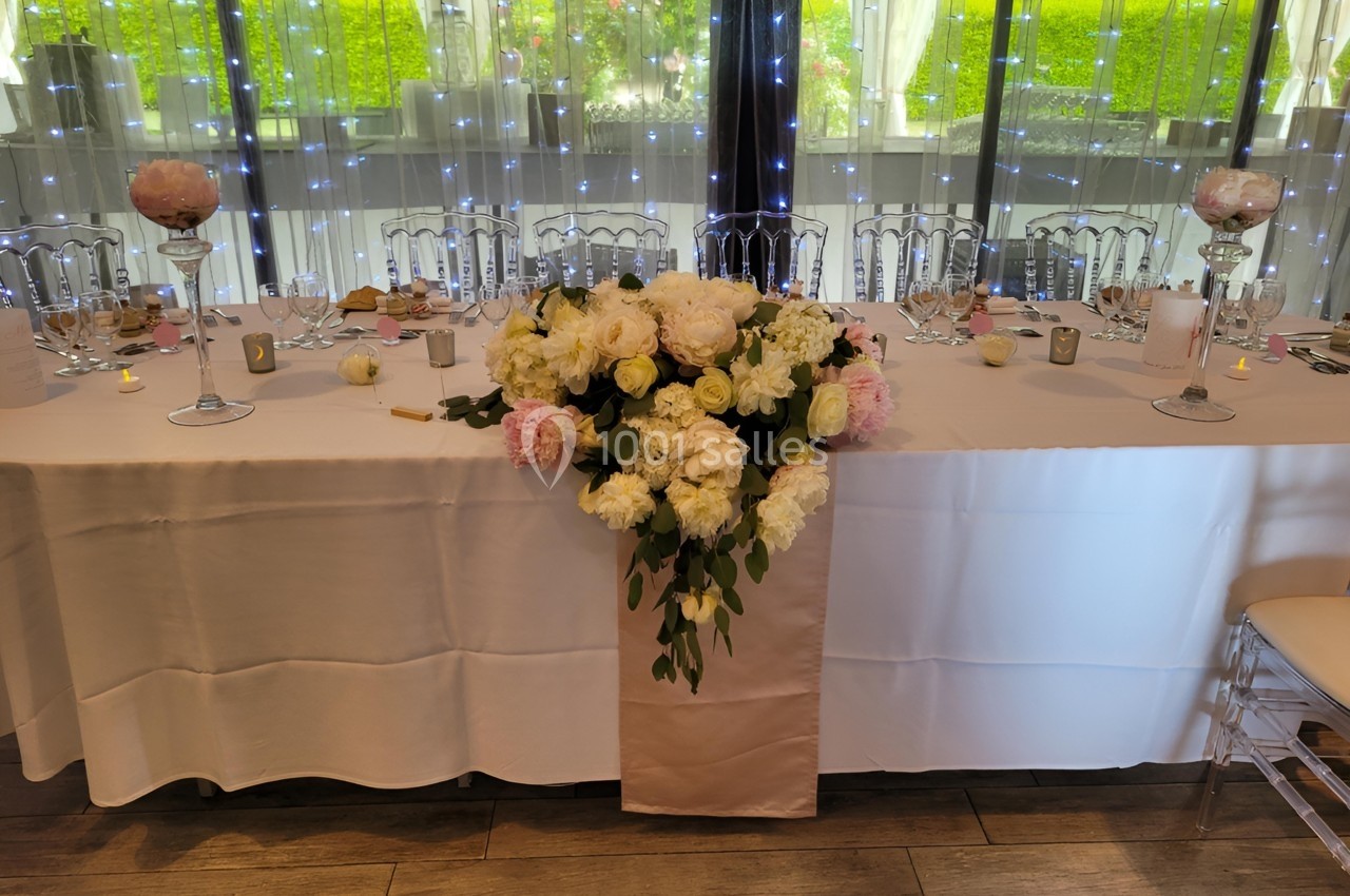 Table de réception décorée avec une nappe blanche, un centre de table floral et des chaises transparentes devant une baie…
