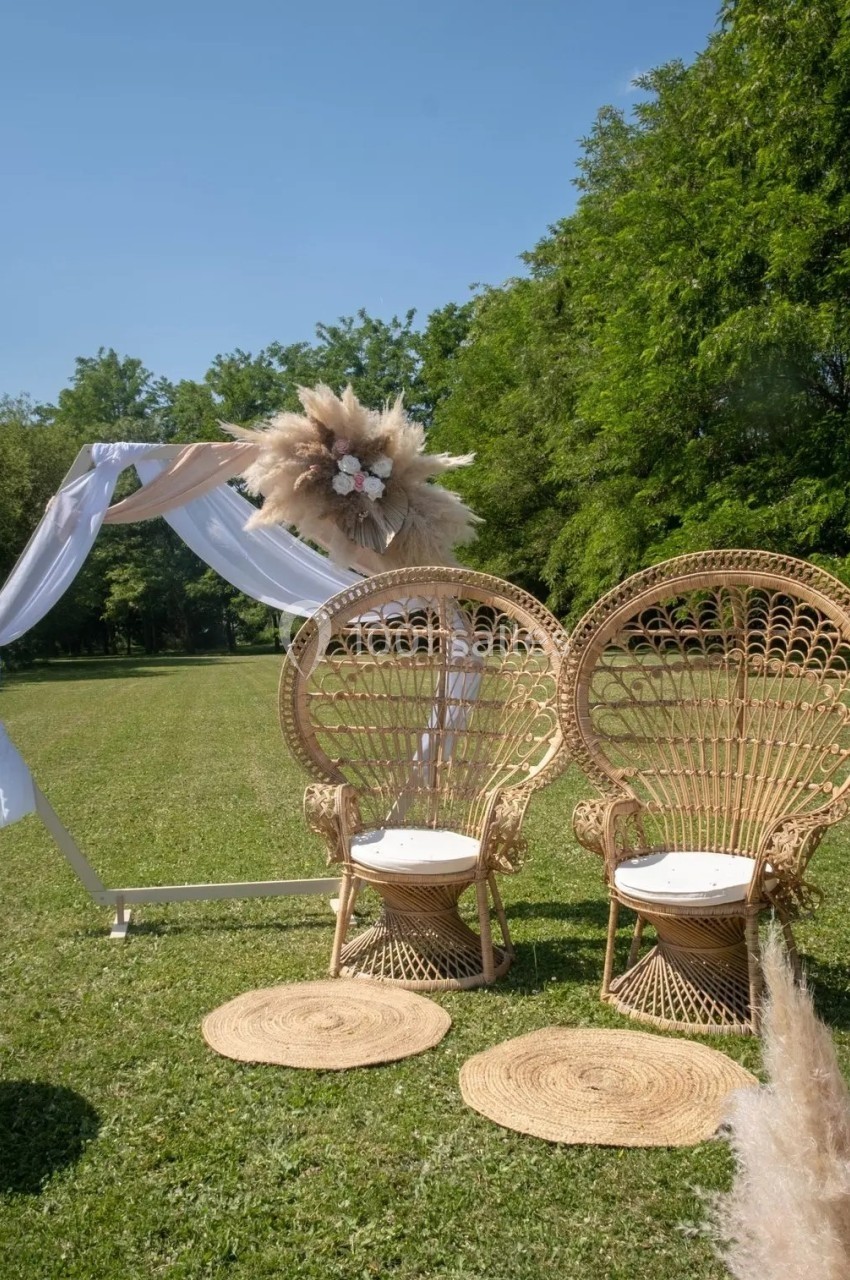 Deux fauteuils en rotin et une arche décorée de fleurs et tissu blanc sur une pelouse entourée d'arbres.