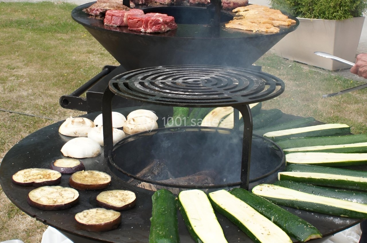 Barbecue circulaire avec légumes grillés (courgettes, aubergines, champignons) et viandes en cuisson sur deux niveaux.