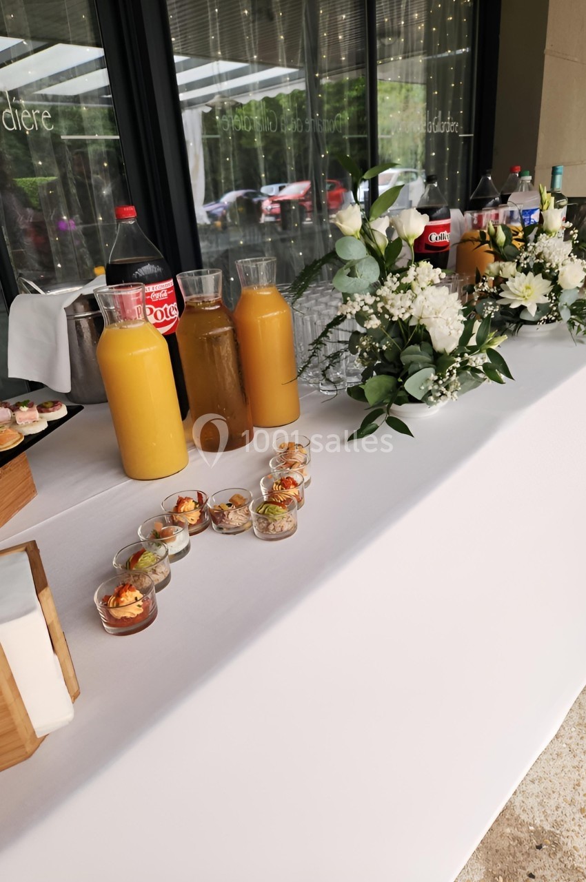 Table de buffet avec boissons, verrines, cupcakes et décoration florale, disposée en extérieur sous un auvent.