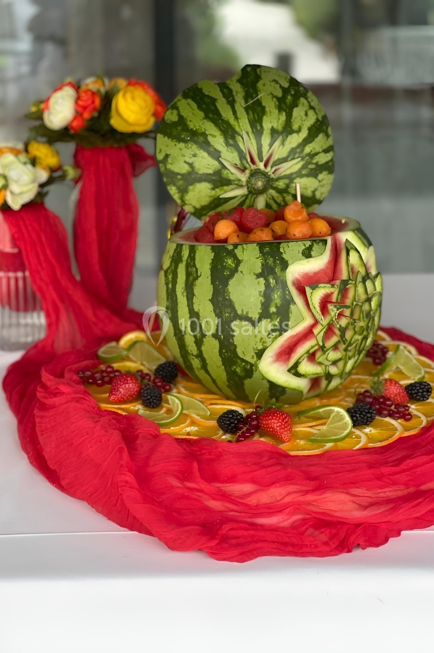 Saladier sculpté dans une pastèque, rempli de fruits variés, entouré de tranches de citron et de fruits rouges.