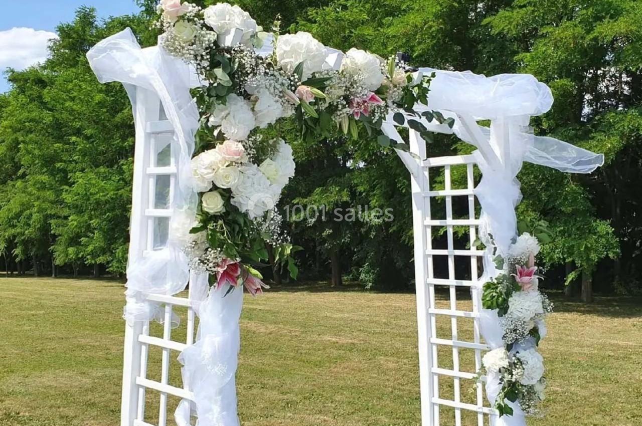 Arche blanche décorée de fleurs blanches et roses avec des voilages, installée dans un parc verdoyant.