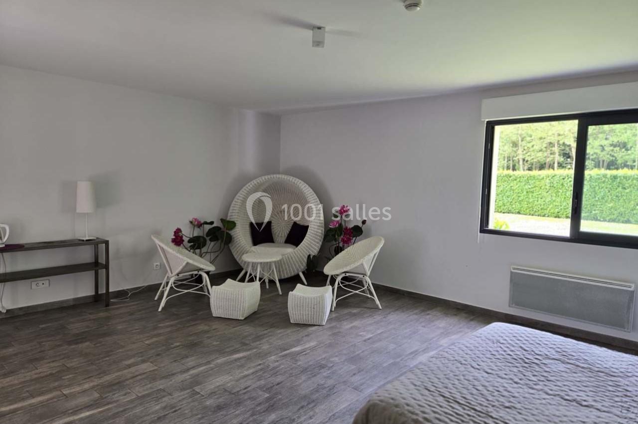 Espace intérieur lumineux avec fauteuils en rotin blanc, table basse, lit et grande fenêtre donnant sur un jardin.