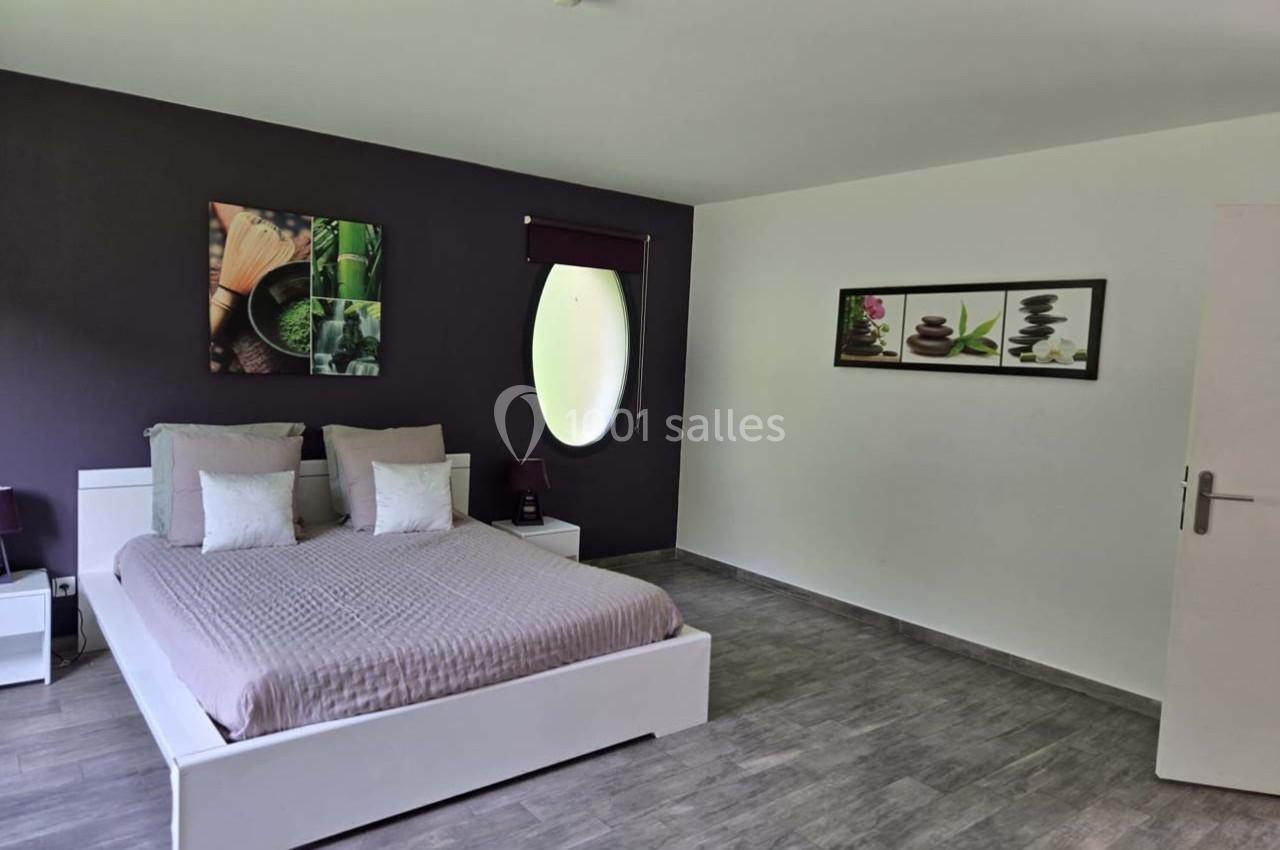 Chambre moderne avec lit double, mur violet décoré de tableaux, sol en parquet gris et fenêtre ovale.