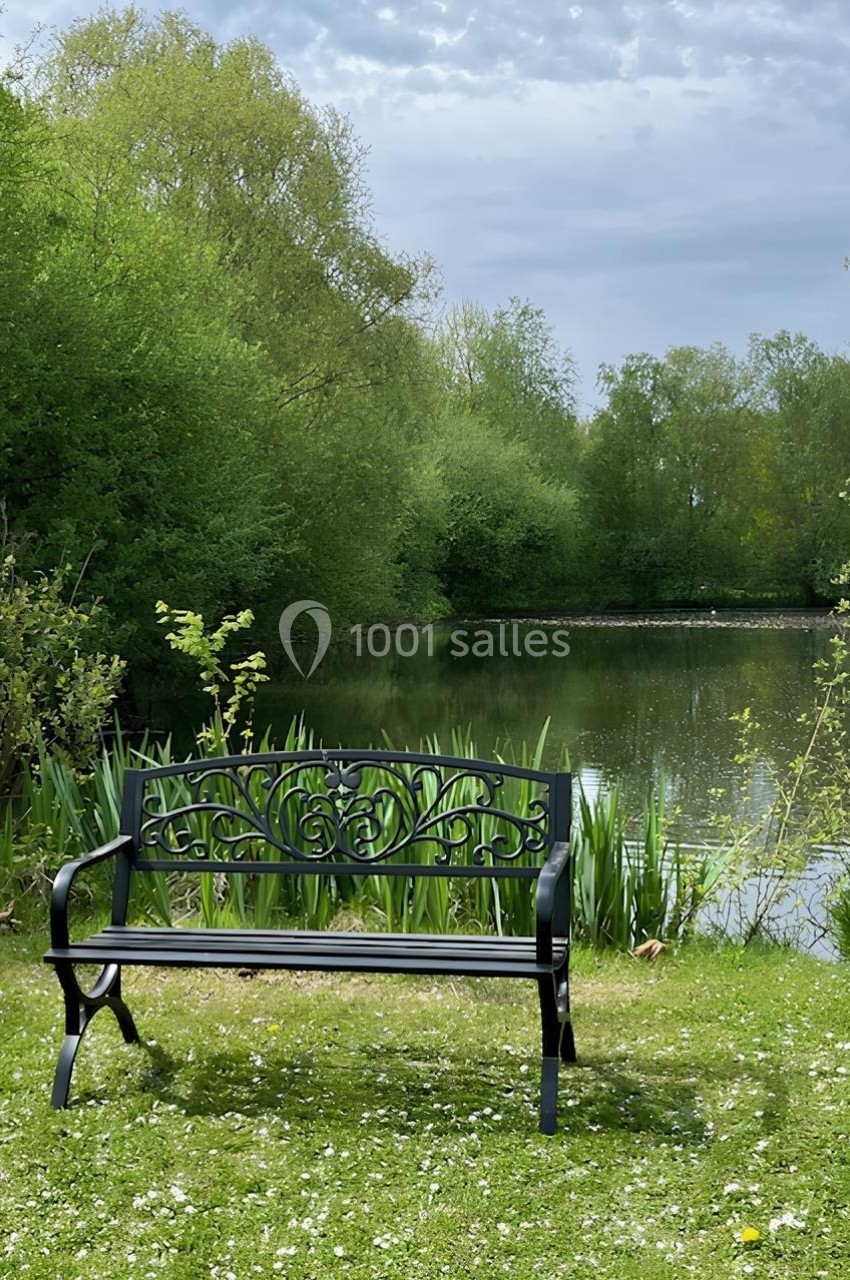 Banc en métal noir placé sur une pelouse, devant un étang entouré de végétation verdoyante sous un ciel nuageux.