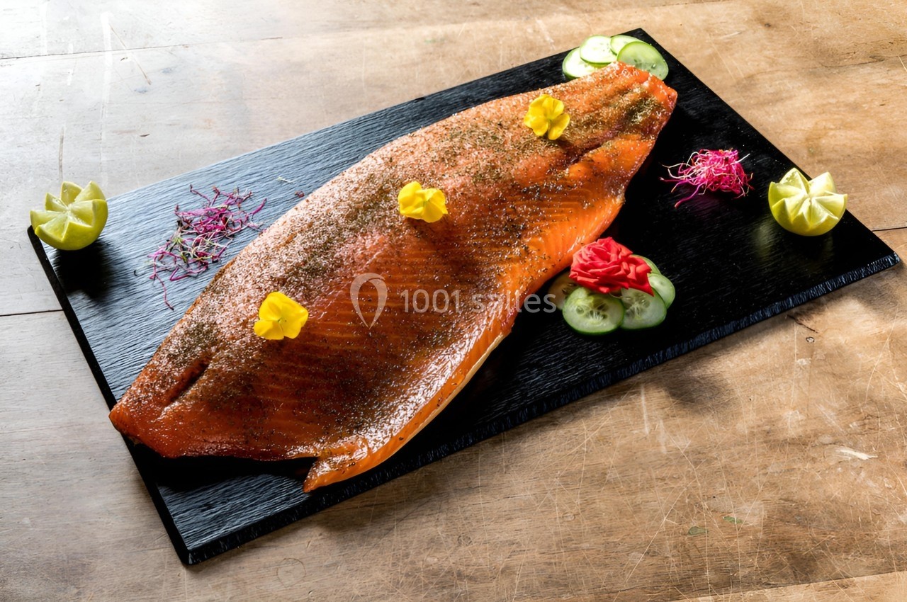 Filet de saumon fumé présenté sur une planche noire, décoré de fleurs comestibles, rondelles de concombre et quartiers de…
