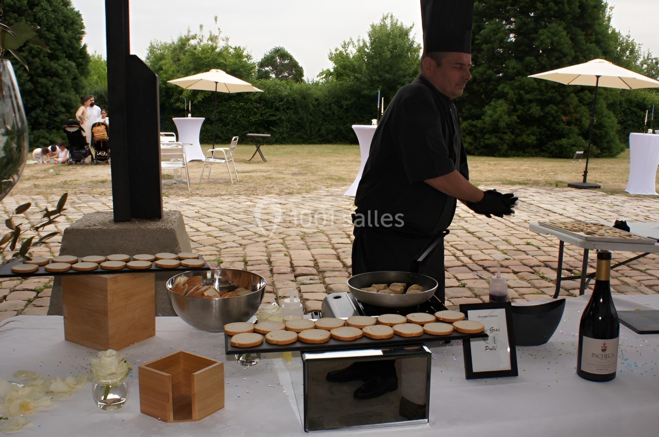 Un chef prépare des amuse-bouches sur une table dressée en extérieur, avec des parasols et un jardin en arrière-plan.