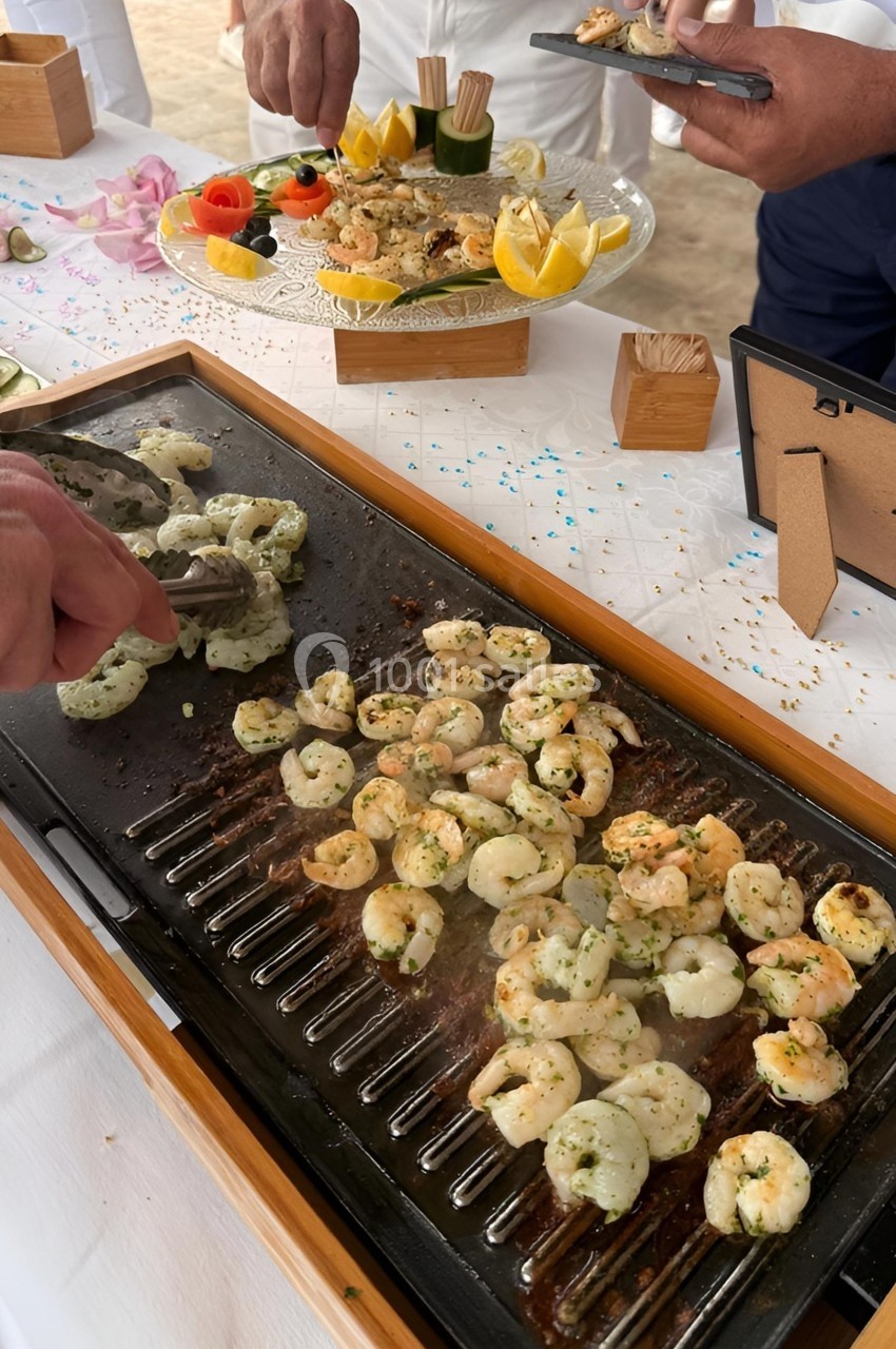 Des crevettes assaisonnées sont grillées sur une plancha, entourées de mains préparant des plats sur une table dressée.