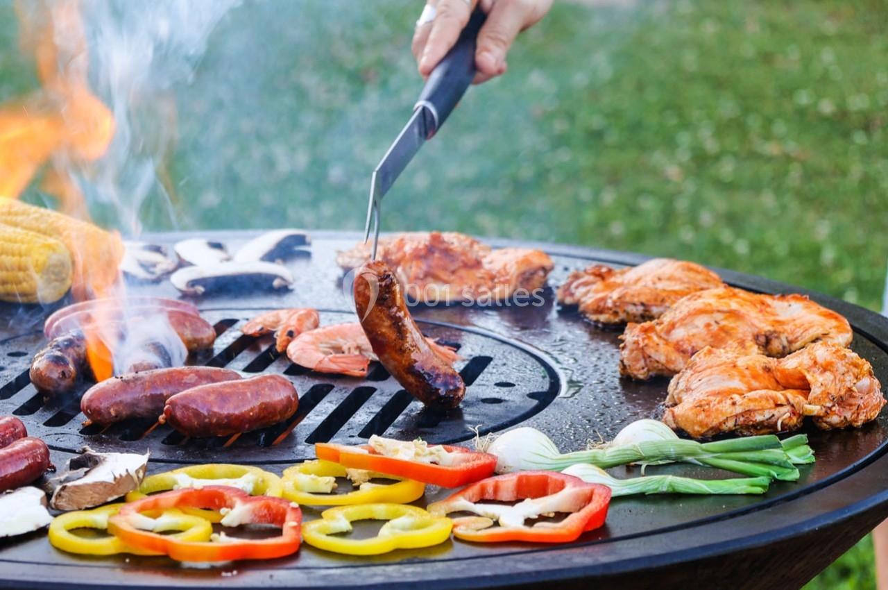 Viande, légumes et épis de maïs grillent sur un barbecue rond en plein air, une main retourne une saucisse.
