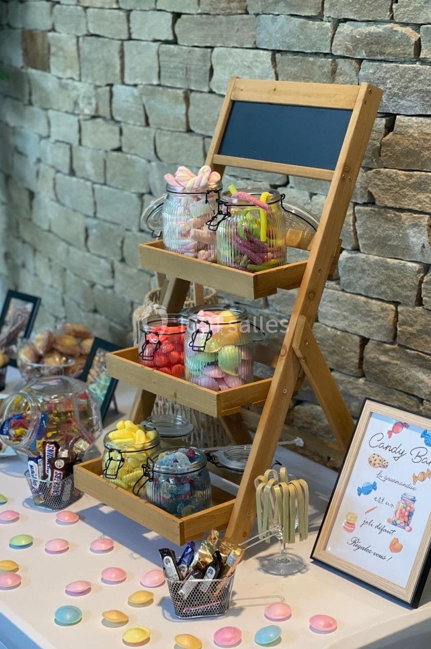 Présentoir en bois avec des bocaux de bonbons colorés, entouré d'autres friandises sur une table blanche.