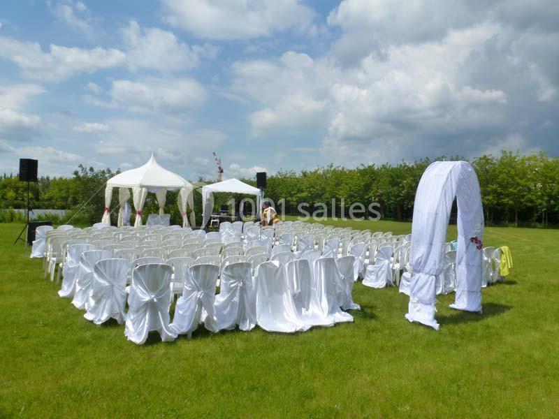 Chaises blanches alignées devant une arche et des tentes dans un jardin, préparées pour une cérémonie en plein air.