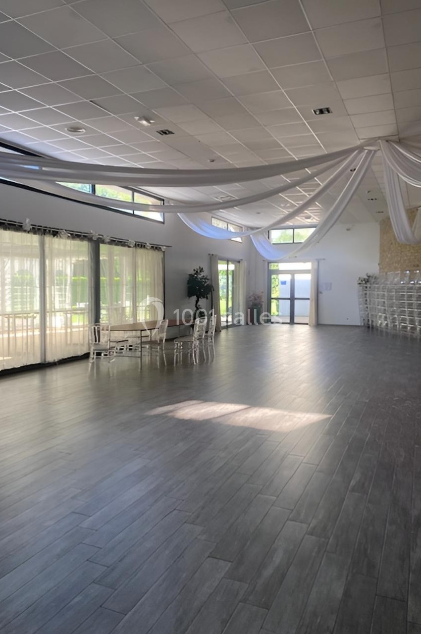 Salle spacieuse avec parquet sombre, grandes baies vitrées, plafond décoré de voilages blancs et chaises empilées.