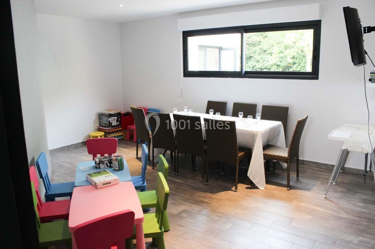 Salle lumineuse avec une table dressée, des chaises en osier, des petites tables colorées et un coin jeux pour enfants.