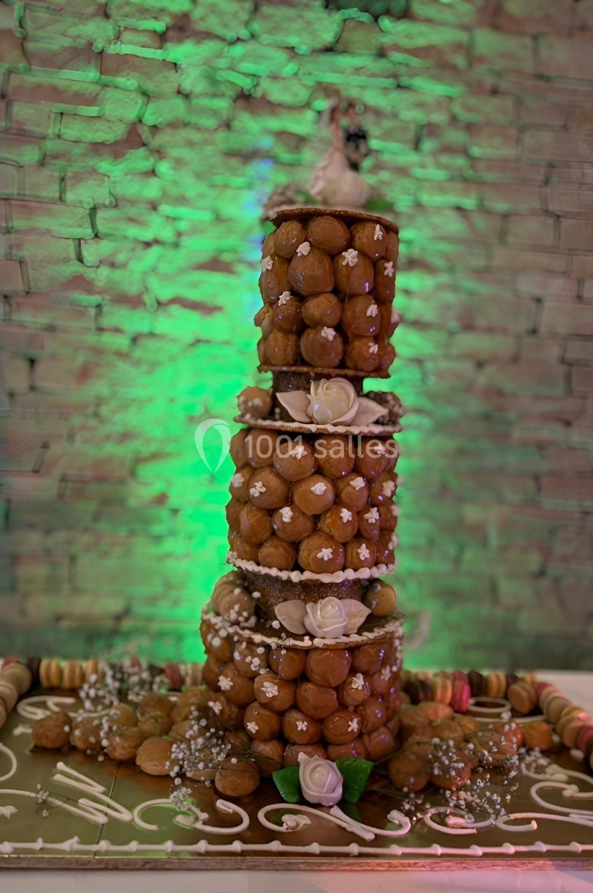 Tour de choux décorée de fleurs et perles, posée sur un plateau orné de macarons, devant un mur en pierre éclairé.