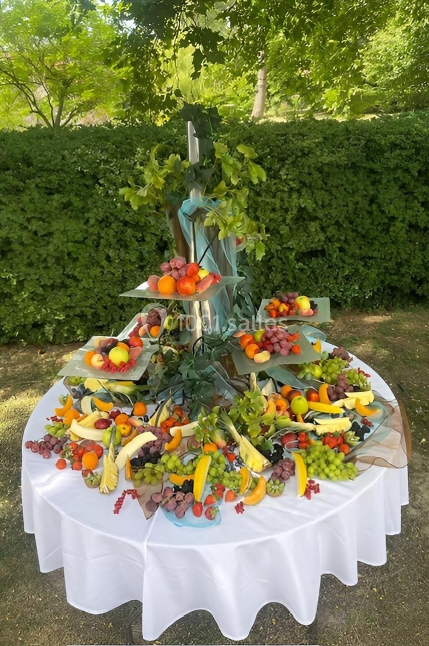 Présentoir de fruits variés disposés sur plusieurs niveaux, entouré de feuillage, sur une table blanche en extérieur.