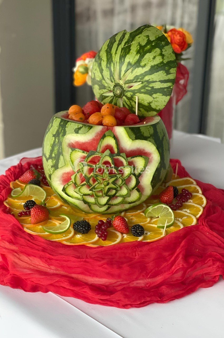 Pastèque sculptée en forme de fleur, remplie de fruits, posée sur un tissu rouge avec des tranches d'agrumes et des baies.
