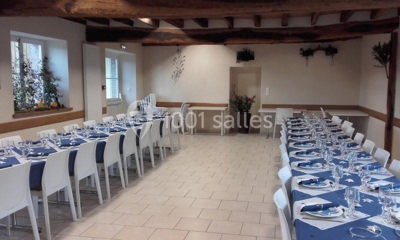 Salle de réception avec tables dressées, nappes bleues, chaises blanches et poutres apparentes au plafond.