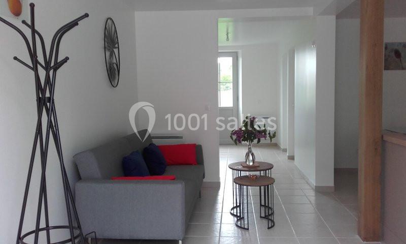 Salon lumineux avec canapé gris, coussins colorés, tables basses et vue sur un couloir menant à une porte-fenêtre.