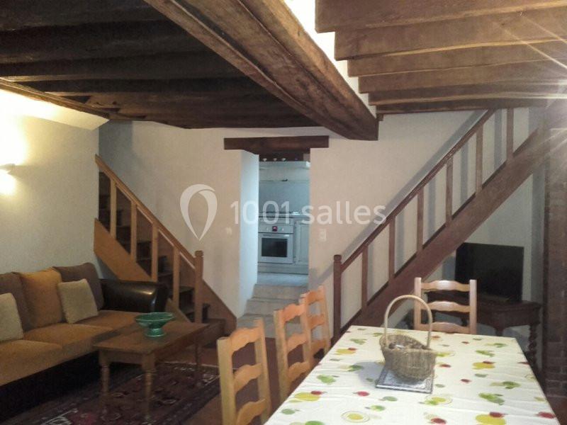 Salon rustique avec poutres apparentes, canapé, table à manger, escalier en bois et vue sur une cuisine ouverte.
