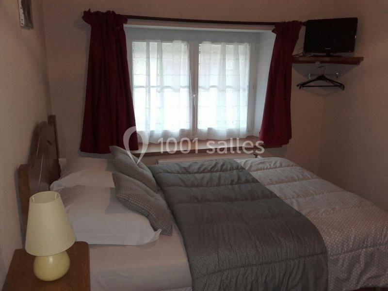 Chambre avec un lit double, des draps gris, des rideaux rouges, une lampe de chevet et une petite télévision murale.