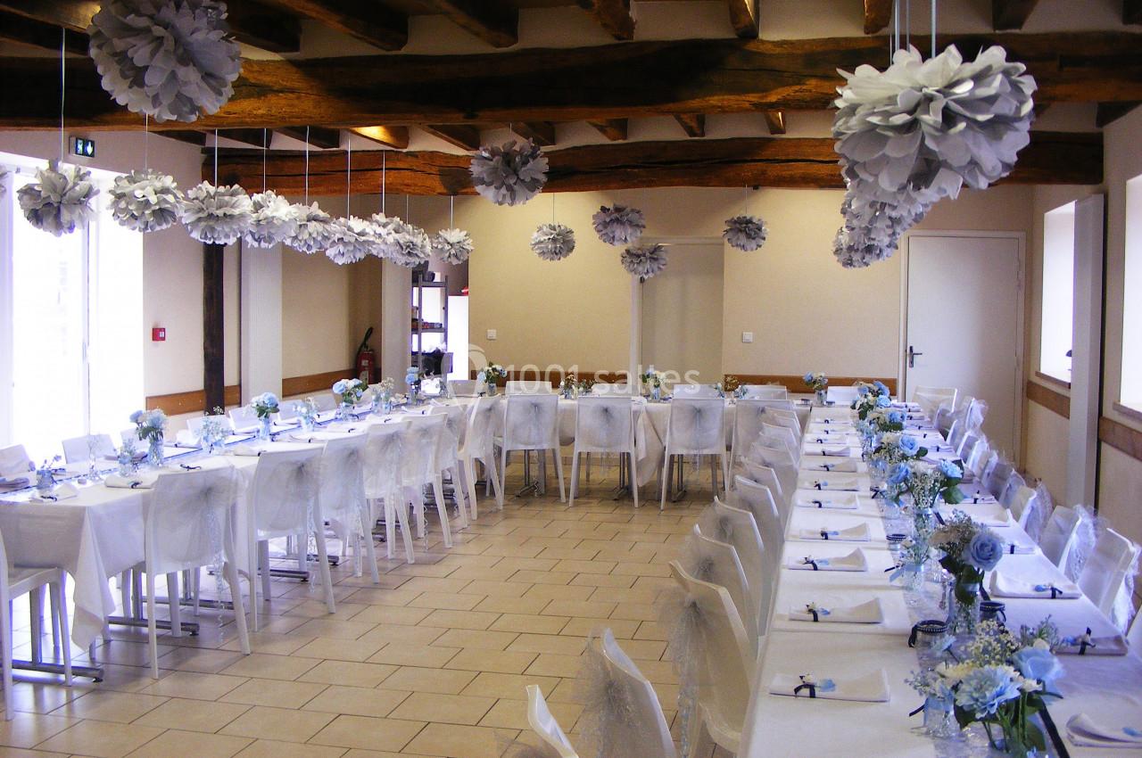 Salle décorée pour un événement, avec tables alignées, nappes blanches, fleurs bleues et suspensions en papier.