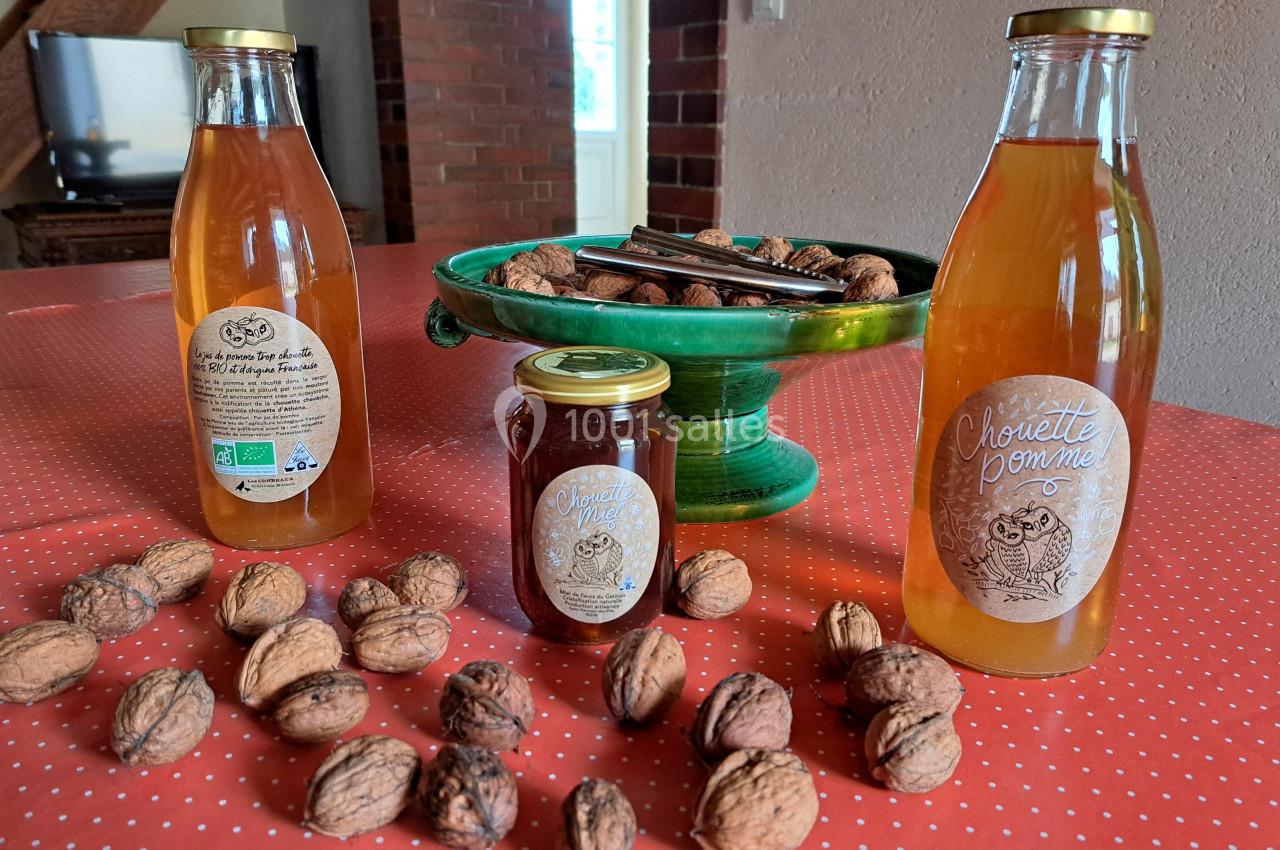 Bouteilles de jus de pomme, pot de confiture et noix disposés sur une nappe rouge à pois, avec un plat vert en arrière-plan.