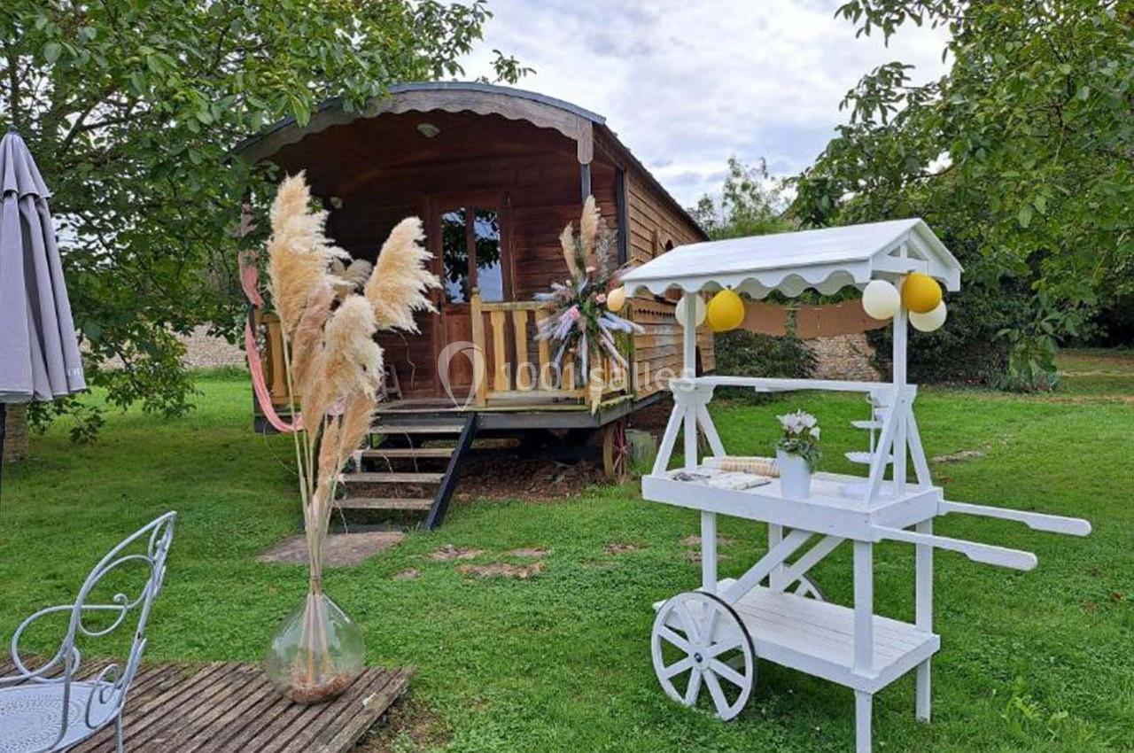 Roulotte en bois dans un jardin verdoyant avec une charrette blanche décorée et des herbes de la pampa en vase.