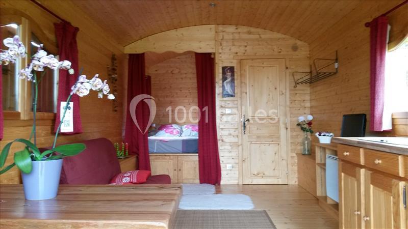 Intérieur d'une roulotte en bois avec un coin salon, un lit en alcôve, une kitchenette et des rideaux rouges.