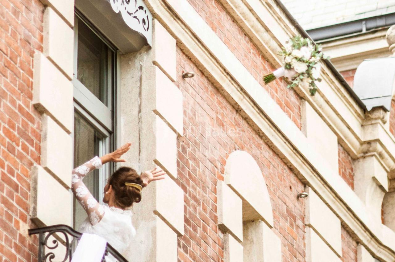 Femme en robe blanche saluant depuis un balcon décoré de tissu et de verdure sur une façade en briques.