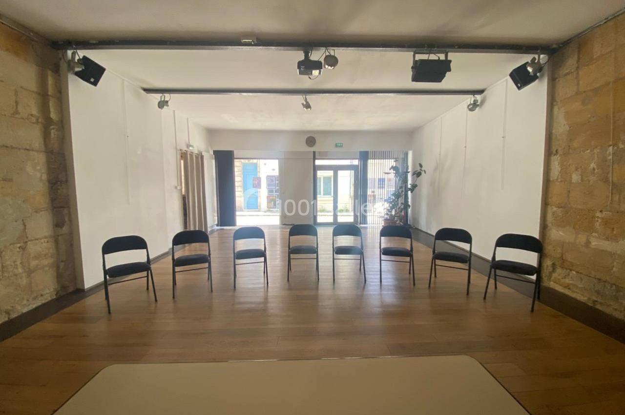 Salle lumineuse avec parquet en bois, murs clairs, chaises noires alignées et grande baie vitrée en arrière-plan.