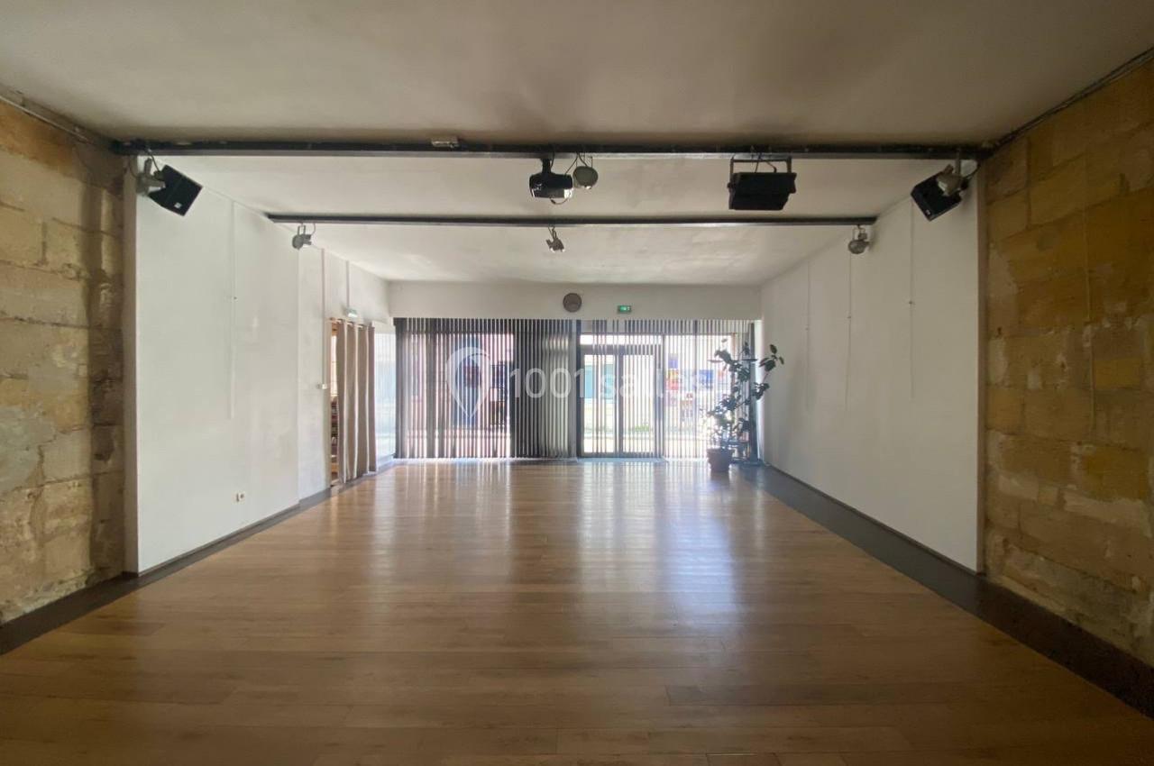 Salle vide avec parquet en bois clair, murs en pierre et en blanc, éclairage au plafond et grande baie vitrée au fond.