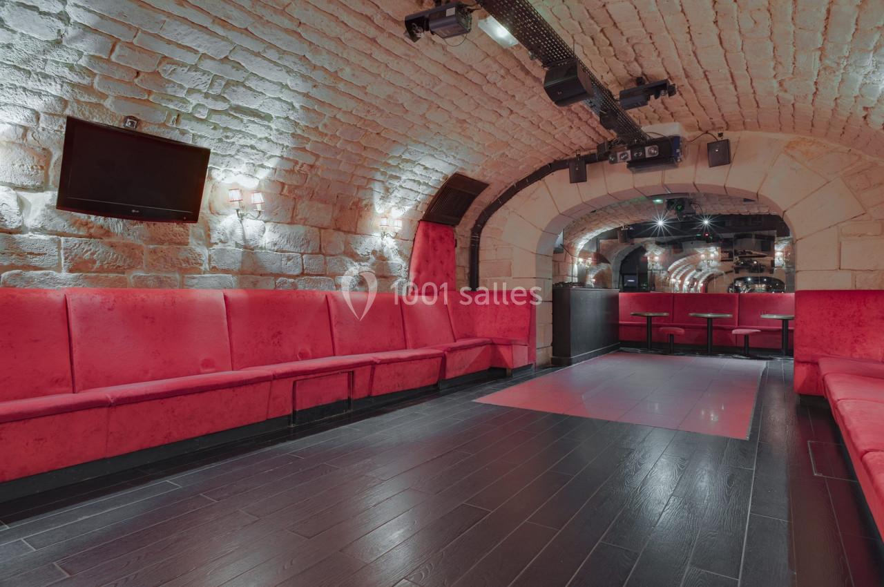 Salle voûtée en pierre avec banquettes rouges, sol en bois sombre et éclairage tamisé.