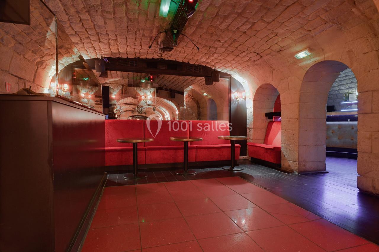 Salle voûtée en pierre avec banquettes rouges, tables basses et éclairage tamisé.