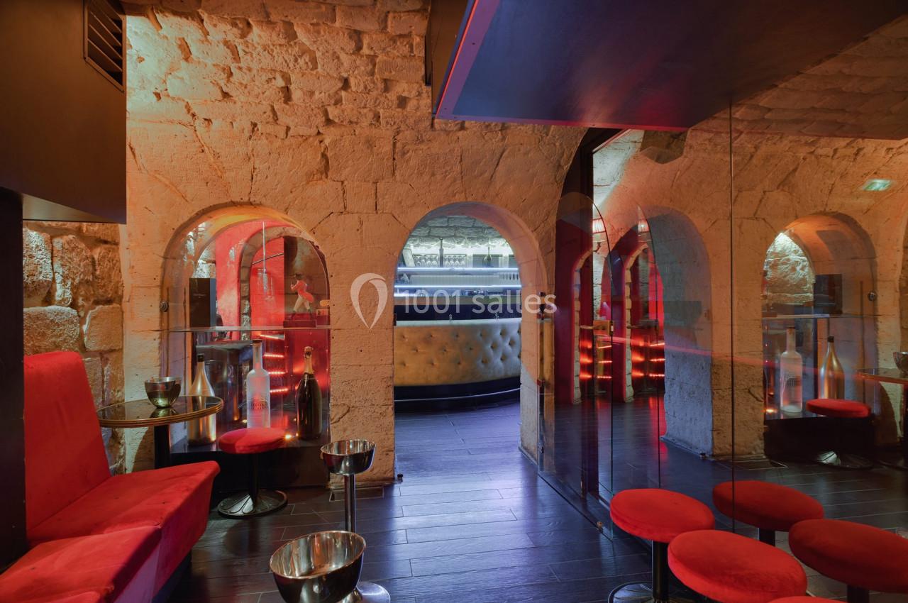 Salle voûtée avec murs en pierre, banquettes rouges, tabourets, et éclairage tamisé près d'un bar moderne.