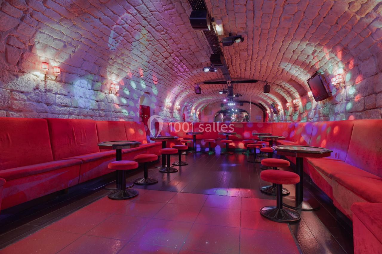 Salle voûtée en pierre avec banquettes rouges, tables rondes et éclairage coloré diffus.