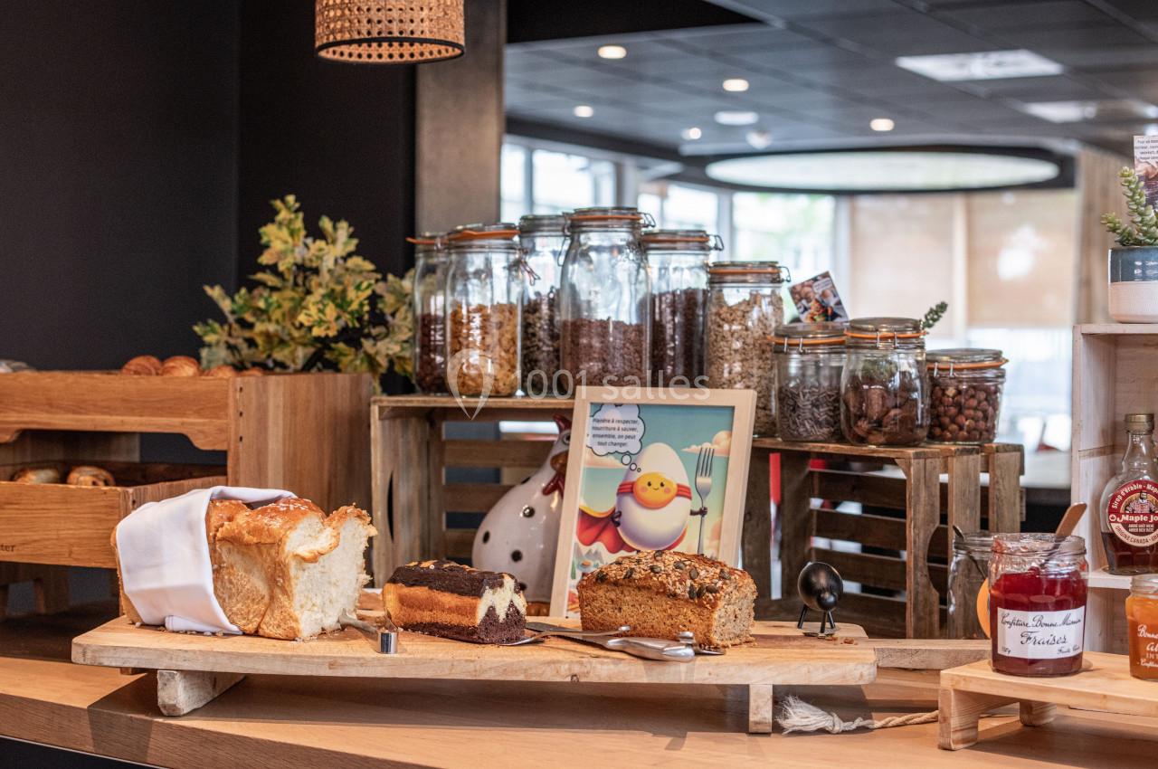 Buffet avec pains, gâteaux, confitures et bocaux de céréales sur une table en bois dans un espace lumineux.