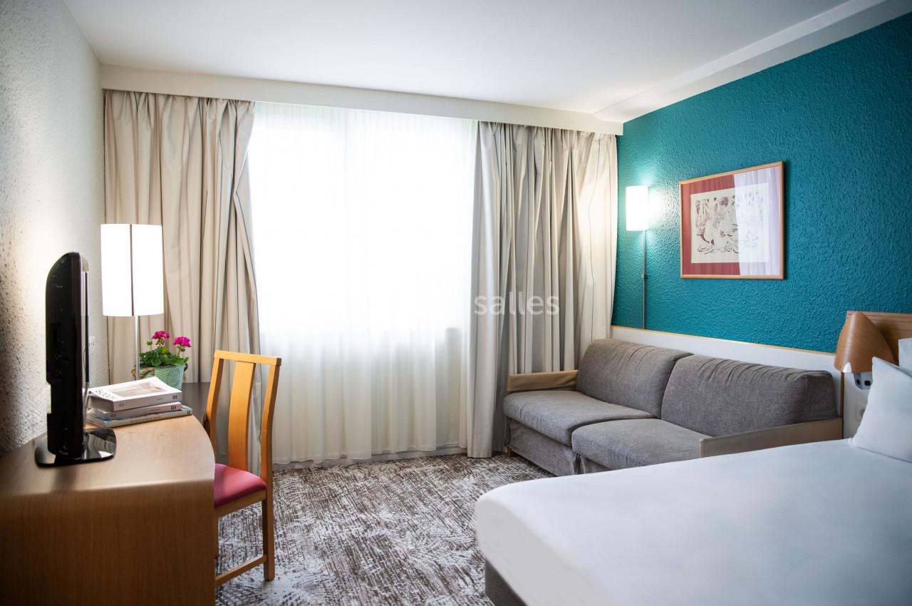 Chambre lumineuse avec un lit, un canapé, un bureau, une télévision et une grande fenêtre avec rideaux.