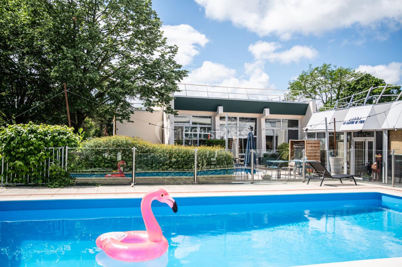 Piscine extérieure avec un flamant rose gonflable, entourée de verdure et d'un bâtiment moderne en arrière-plan.