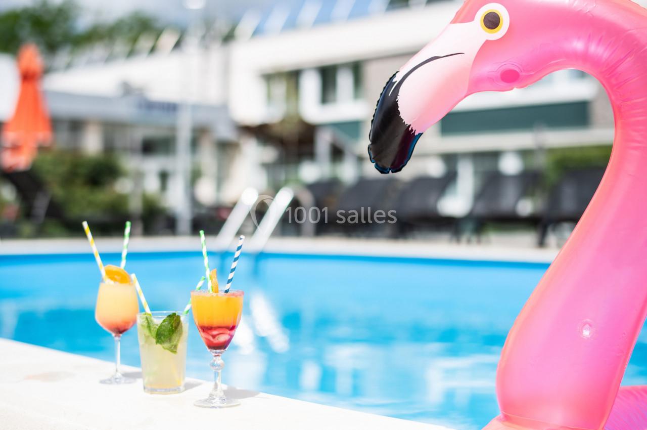 Gros plan sur un flamant rose gonflable et trois cocktails colorés au bord d'une piscine extérieure.