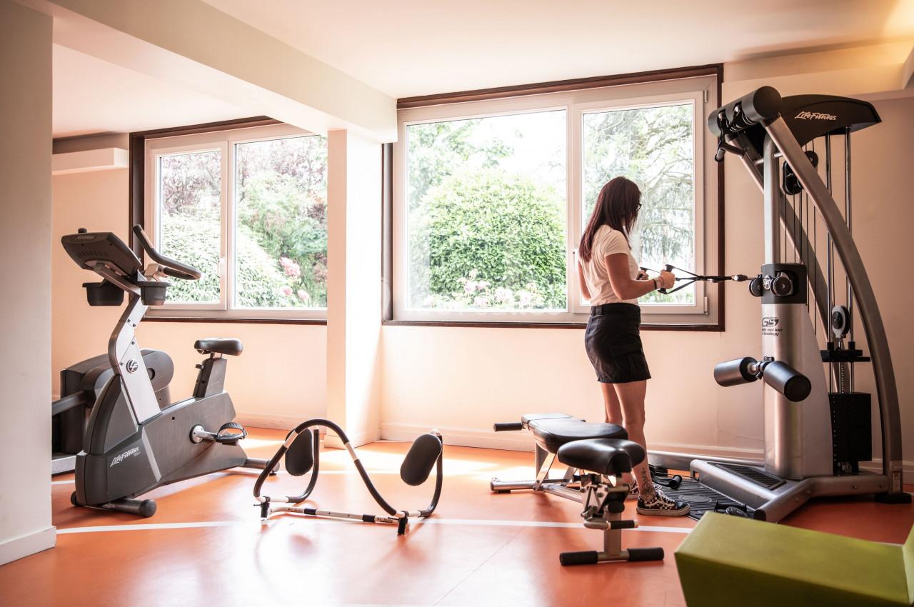 Femme utilisant une machine de musculation dans une salle de sport lumineuse avec vue sur un jardin.