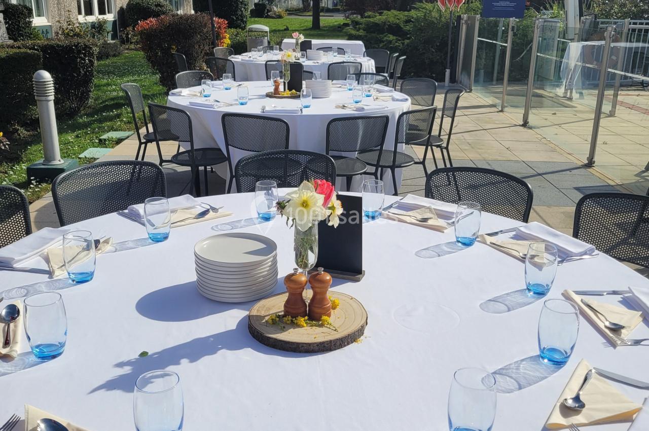 Tables dressées en extérieur avec nappes blanches, verres bleus et décoration florale, sous un ciel ensoleillé.