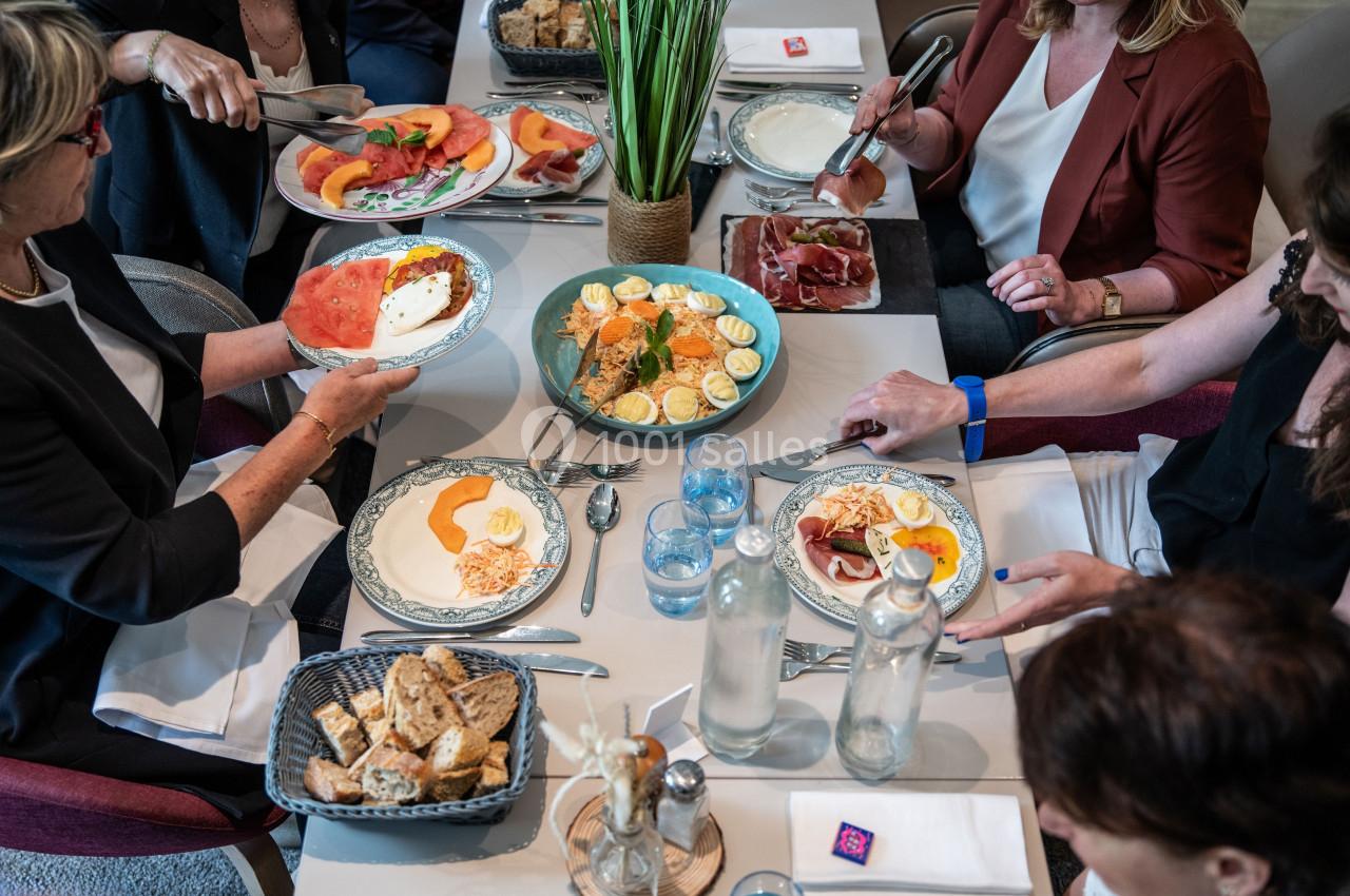 Un groupe de personnes partage un repas composé de fruits, œufs, charcuterie et pain autour d'une table dressée.