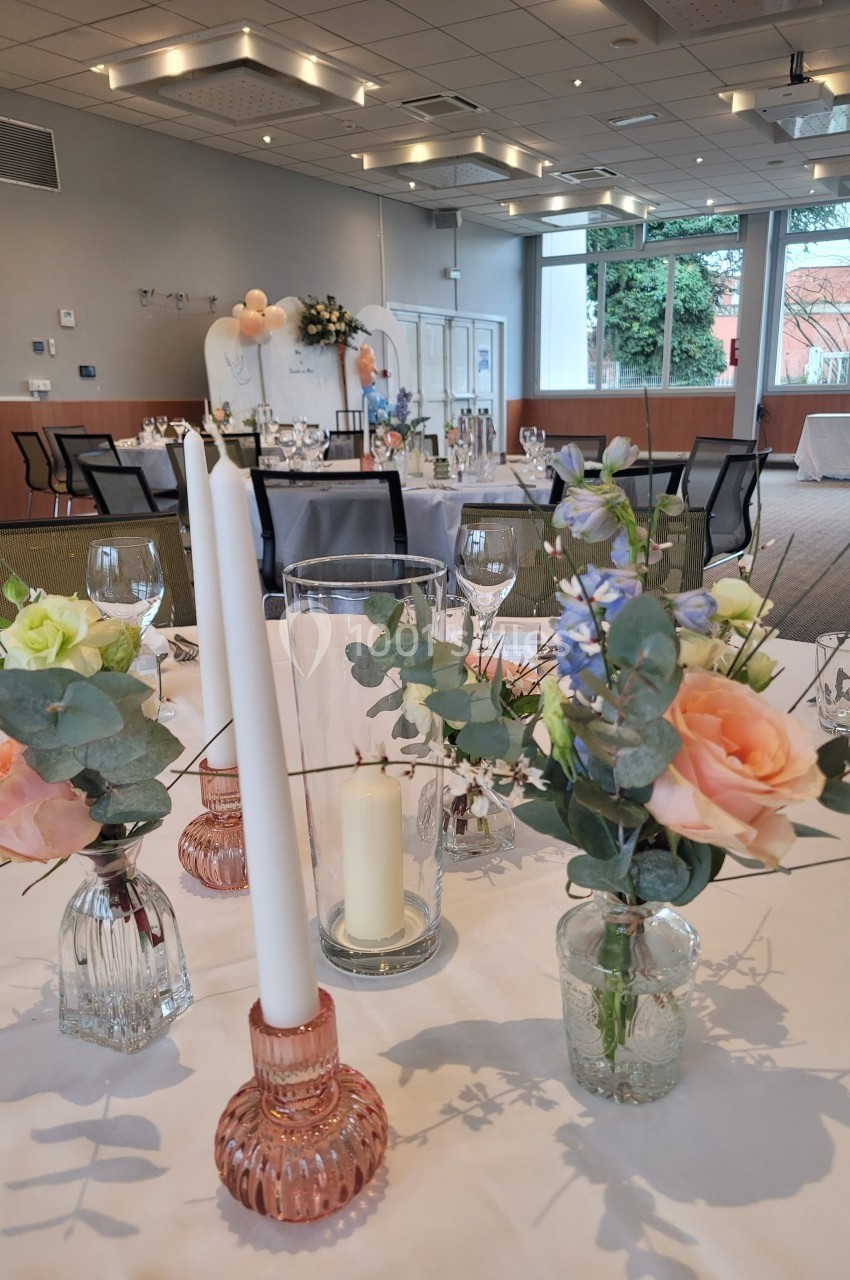 Décoration de table avec bougies, fleurs pastel et verrerie dans une salle lumineuse préparée pour un événement.
