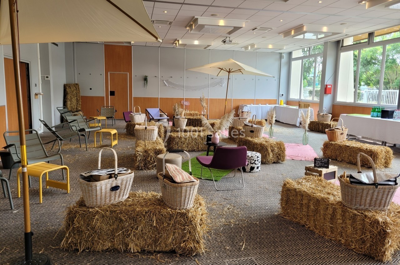 Salle aménagée avec des bottes de paille, des parasols, des chaises et des paniers, dans un cadre lumineux et convivial.