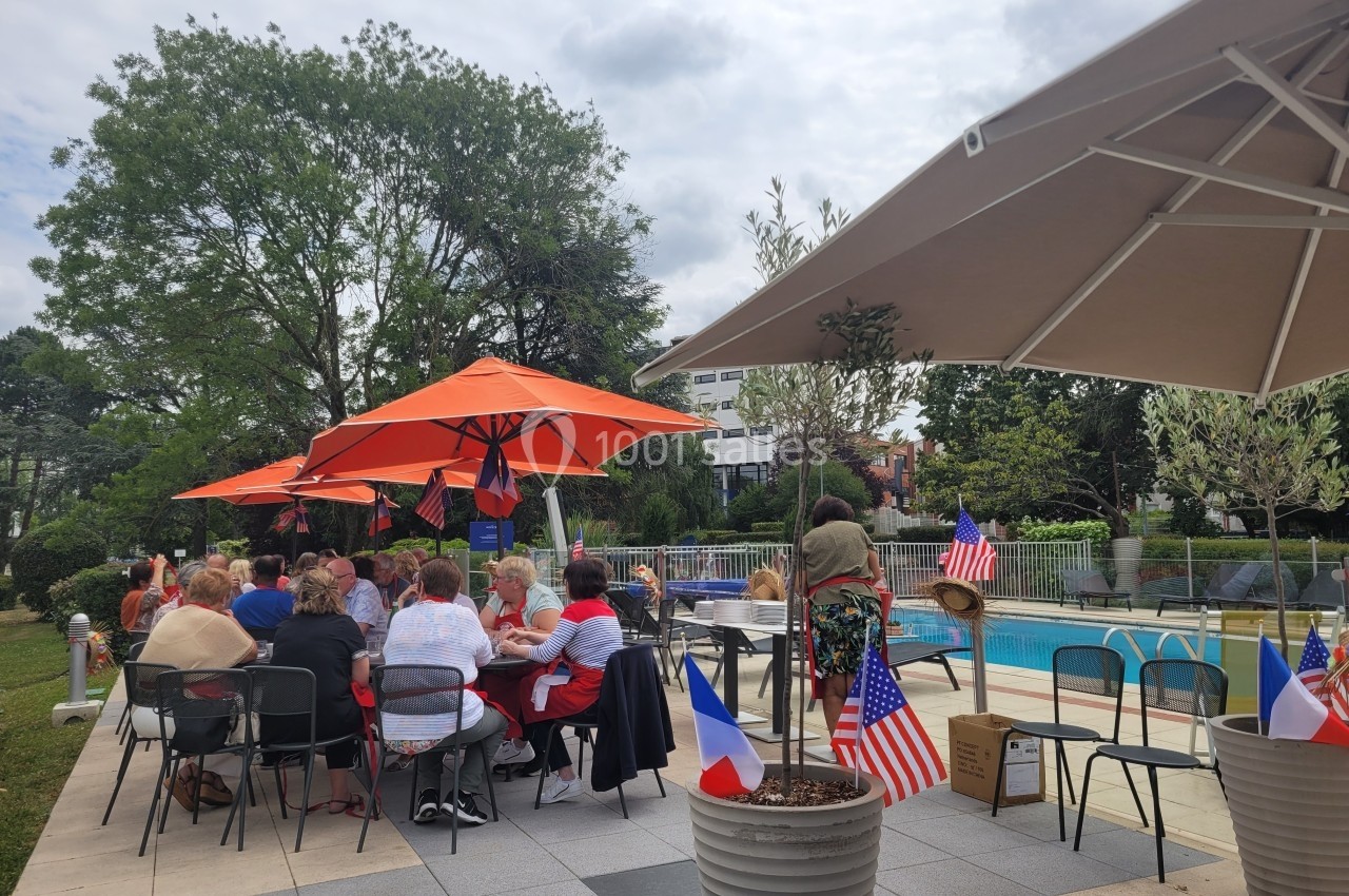 Groupe de personnes assises à des tables en extérieur près d'une piscine, décorées de drapeaux français et américains.