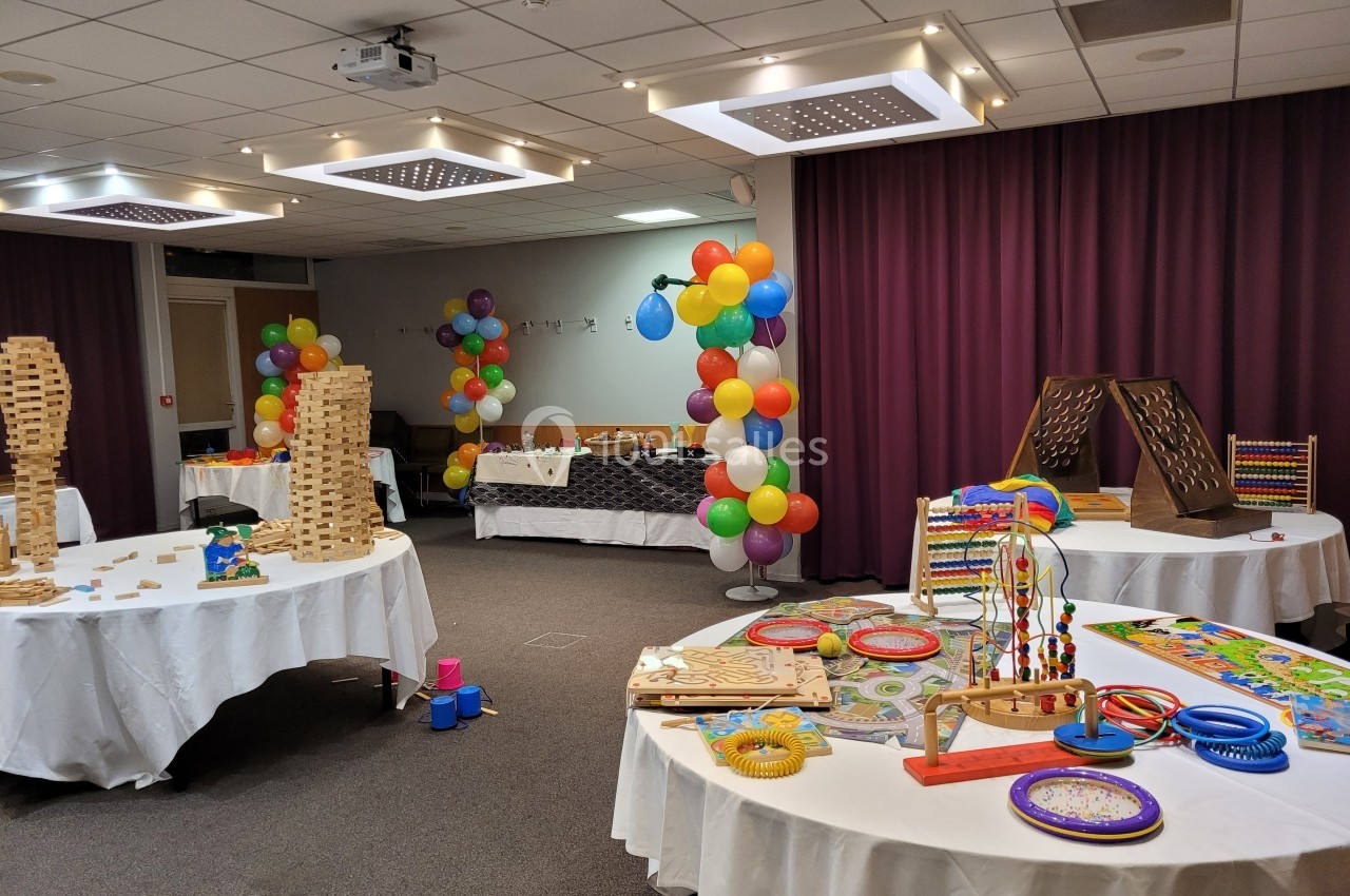 Salle décorée avec des ballons colorés, présentant des tables garnies de jeux et jouets éducatifs en bois.