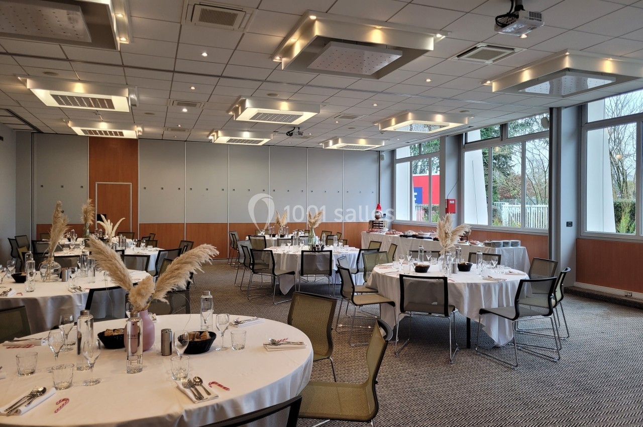 Salle de réception lumineuse avec tables rondes dressées, décorées de plumes blanches, et grandes fenêtres donnant sur l…