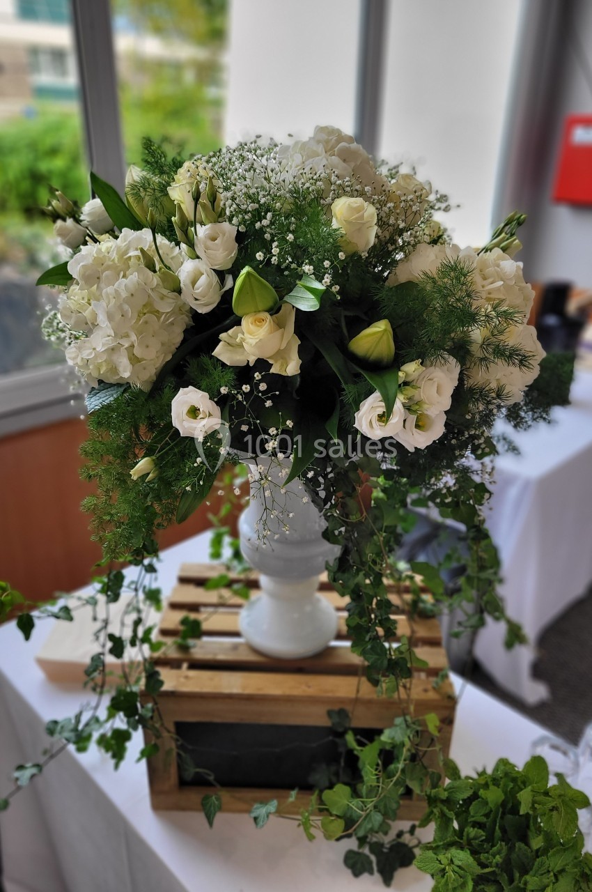Bouquet de fleurs blanches et vertes dans un vase blanc, posé sur une caisse en bois avec des feuillages tombants.